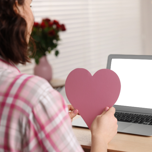 Femme tenant un cœur devant son ordinateur, symbole d’une relation à distance pleine d’affection.