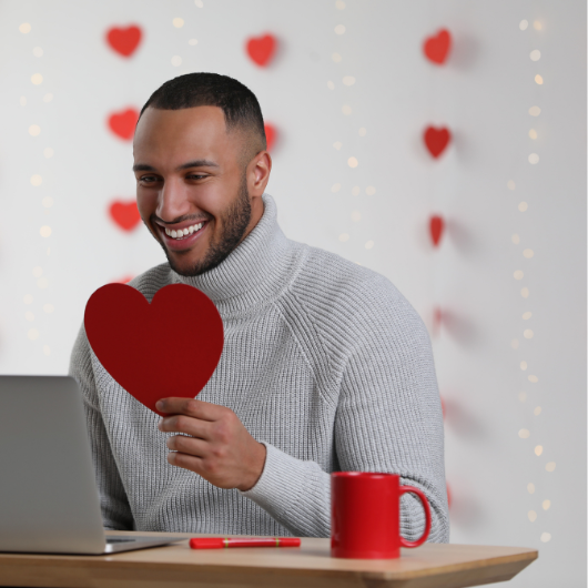 Homme souriant devant un écran, cœur en main, exprimant l’amour malgré la distance physique.
